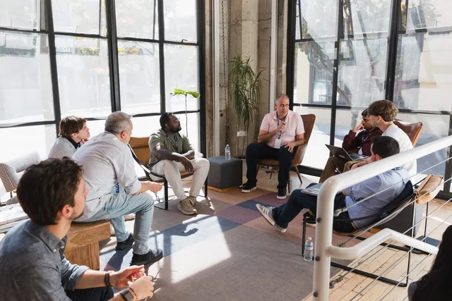 Marlon in a leather armchair in a sunlit loft at Cognition's offices, one hand raised mid-sentence, talking to about eight people seated in a loose circle around him. Tall factory windows and a potted plant stand behind.
