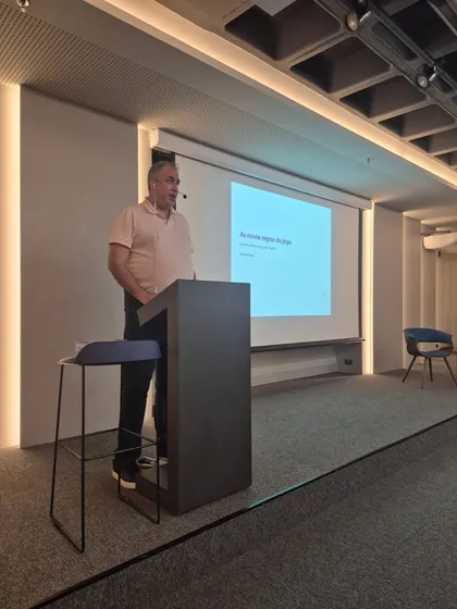 Marlon standing at a dark lectern on a low stage in a modern auditorium, mid-sentence, wearing a headset microphone. On the projection screen behind him: “As novas regras do jogo — Escrever software virou trivial. E agora?”