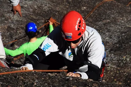 Marlon mid-climb on a rock face, in a red helmet taped with his name and dark sunglasses, one hand reaching for the next hold. A second climber and coils of rope are out of focus behind him.